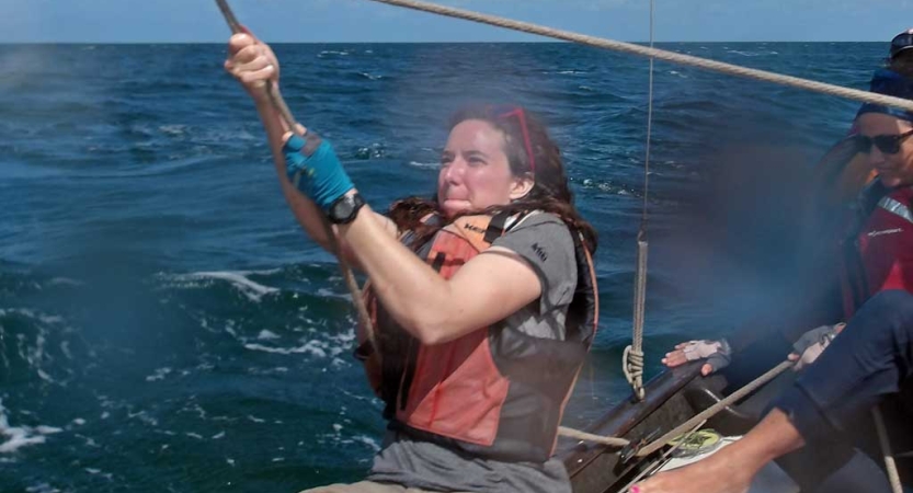 A person wearing a lifejacket sits on a sailboat and pulls on a rope.